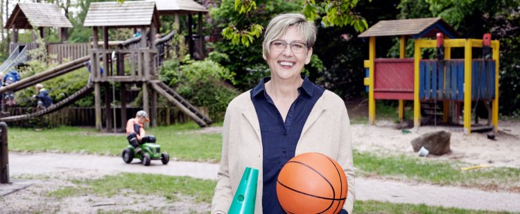 Heilerziehungspflegerin steht auf Spielplatz und hält Kegel und Basketball.