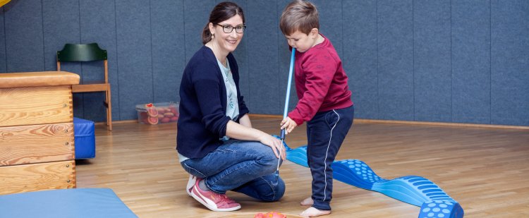 Motopädin kniet in Sporthalle neben einem Jungen, der mit einer Angel Spielzeug vom Boden greift.