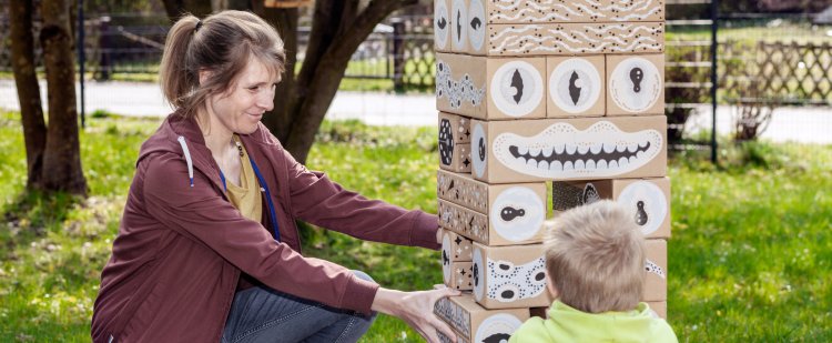 Sozialpädagogin spielt mit Kind im Garten an Turm aus großen Holzklötzen.