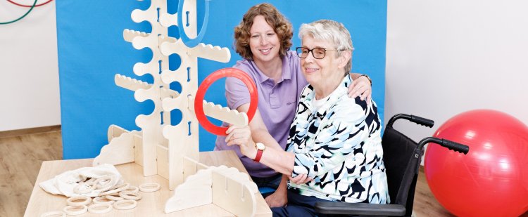 Ergotherapeutin sitzt neben einer Frau im Rollstuhl, die einen roten Ring an eine Holzkonstruktion hängt.