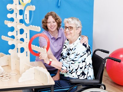 Ergotherapeutin sitzt neben einer Frau im Rollstuhl, die einen roten Ring an eine Holzkonstruktion hängt.