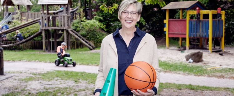 Heilerziehungspflegerin steht auf Spielplatz und hält Kegel und Basketball.