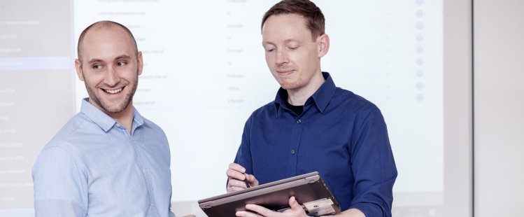 Zwei IT-Fachinformatiker stehen vor Whiteboard mit Handy und Tablet in den Händen.