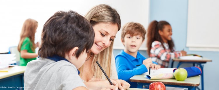 Frau sitzt in Schulklasse neben einem Jungen und hilft ihm bei Aufgaben.