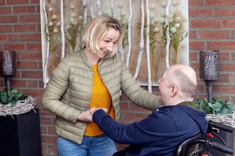 Wohnheimhelferin steht mit Klienten im Rollstuhl vor Blumendeko.