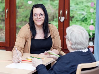 Altenpflegerin und ältere Frau sitzen am Tisch und malen ein Bild.