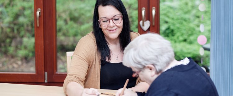 Altenpflegerin und ältere Frau sitzen am Tisch und malen ein Bild.