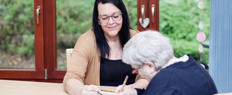 Altenpflegerin und ältere Frau sitzen am Tisch und malen ein Bild.