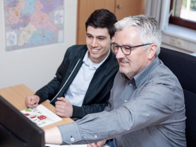 Betriebswirt sitzt neben Azubi am Schreibtisch und zeigt auf einen PC-Bildschirm.