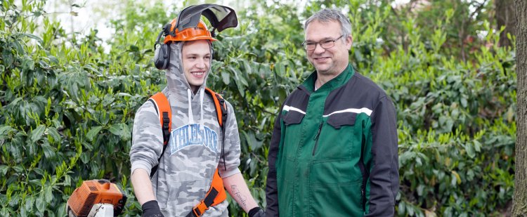 Gärtner und Beschäftigter mit Heckenschneider vor einer Hecke.