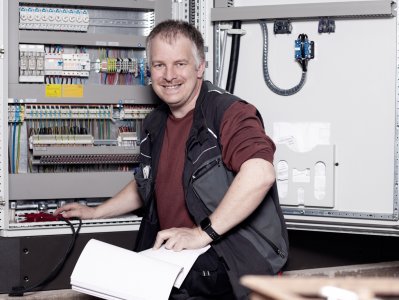 Hausmeister kniet vor einem geöffneten Schaltschrank und hält ein Messgerät hinein.