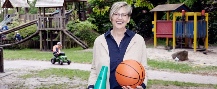 Heilerziehungspflegerin steht auf Spielplatz und hält Kegel und Basketball.