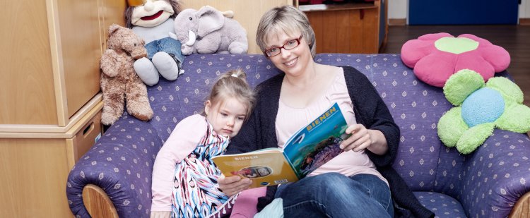 Heilpädagogin sitzt mit Kind auf Sofa und liest aus Buch vor.