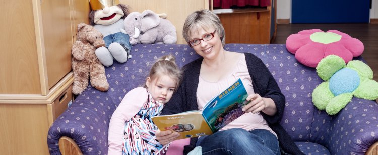 Heilpädagogin sitzt mit Kind auf Sofa und liest aus Buch vor.