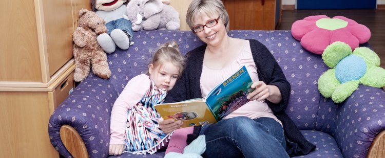 Heilpädagogin sitzt mit Kind auf Sofa und liest aus Buch vor.