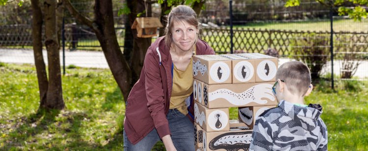 Sozialpädagogin spielt mit Kind im Garten an Turm aus großen Holzklötzen.