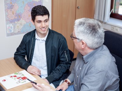 Auszubildener im Büromanagement spricht am Schreibtisch mit Ausbilder.