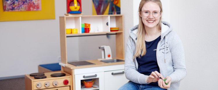 Auszubildende sitzt an einem kleinen Tisch im Kindergarten und spielt mit Knete.