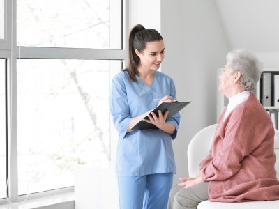 Praktikantin hält Klemmbrett und spricht mit alter Frau, die auf der Bettkante sitzt.