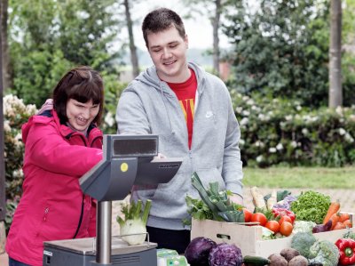 Auszubildender und Klientin stehen draußen hinter einer Theke mit einer Waage und Gemüsekisten.