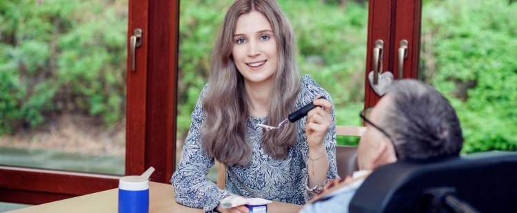 Eine junge Frau gibt einem Menschen im Rollstuhl etwas zu essen.
