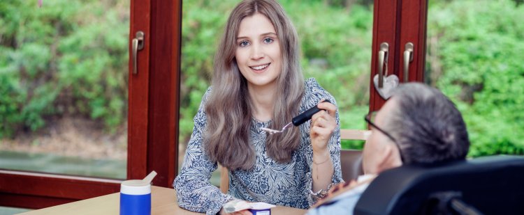 Eine junge Frau gibt einem Menschen im Rollstuhl etwas zu essen.