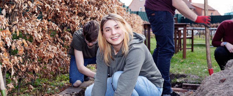 Praktikantin kniet im Beet und macht Gartenarbeit.